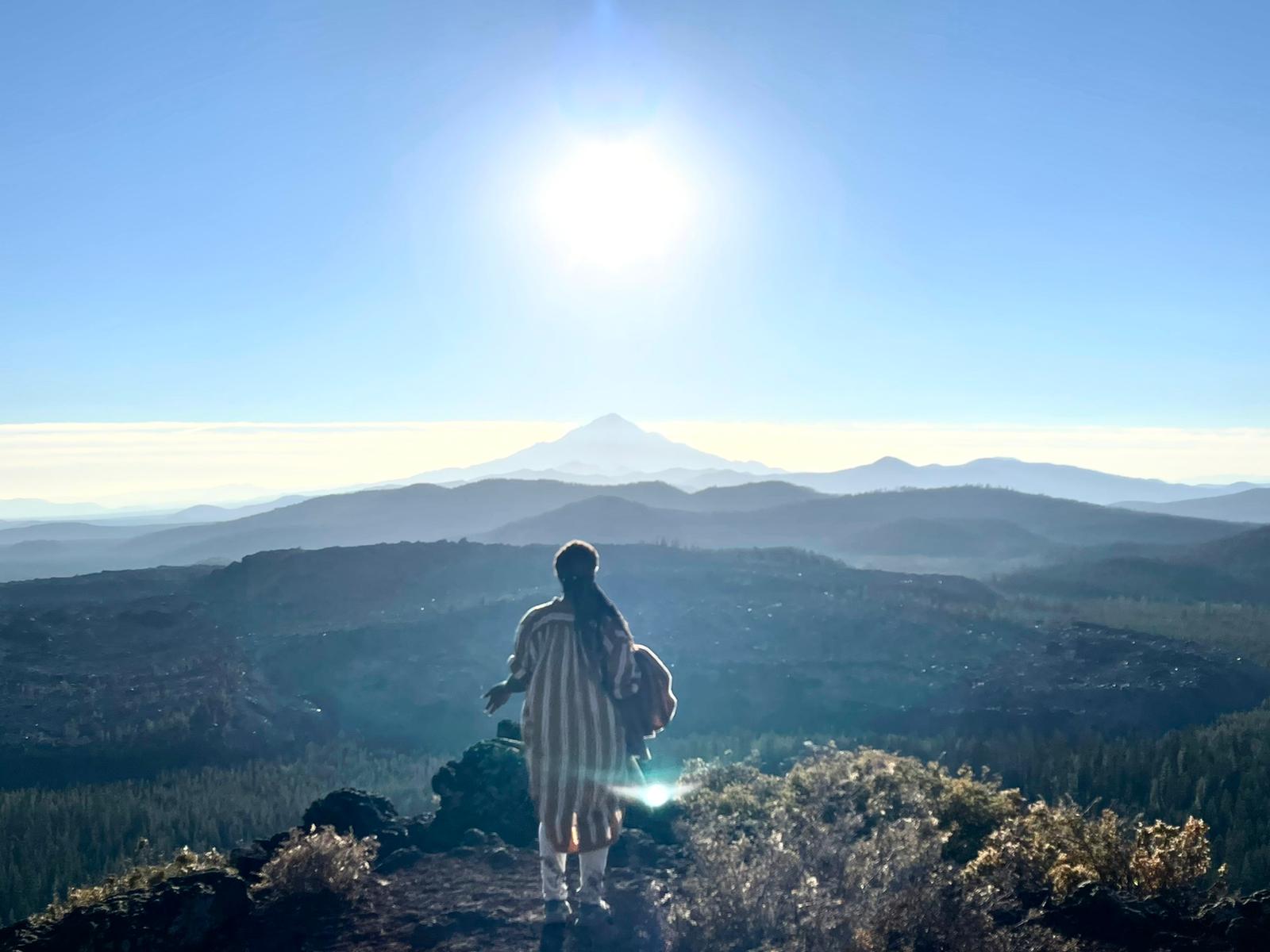 A student standing at a lookout above the valley, facing the sun and the distant mountain